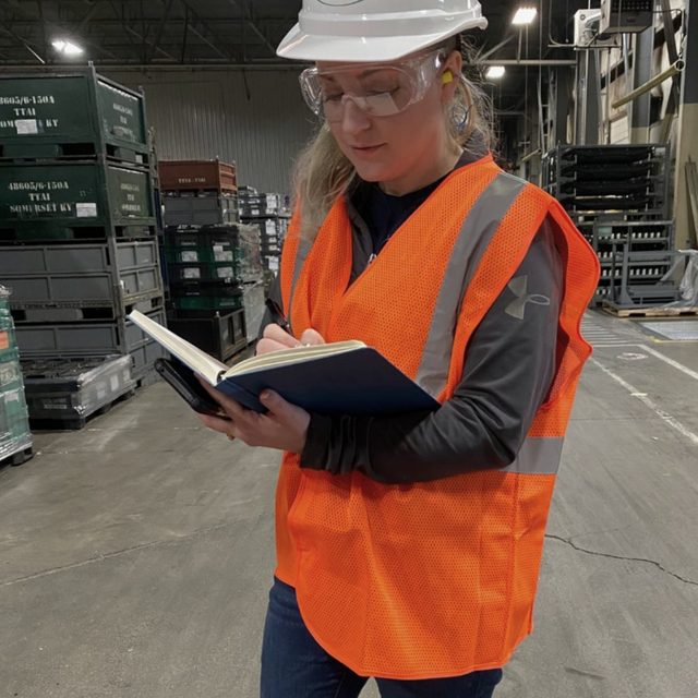 Hollie taking notes on a warehouse floor in a hard hat and safety vest